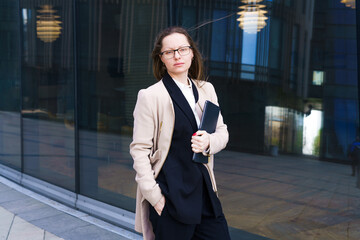 Business woman of Caucasian ethnicity in a coat and suit with a pensive look stands in glasses near the business center. The concept of solving complex business problems