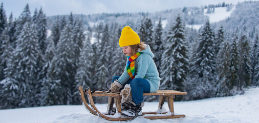 Portrait of happy little kid wearing knitted hat, scarf and sweater. Kid boy enjoying a sleigh ride on winter landscape. Child sledding riding a sleigh outdoors in snow. Winter knitted kids clothes.