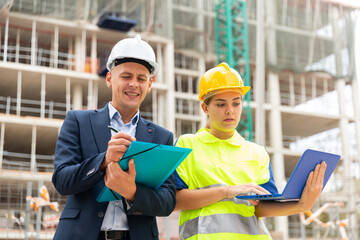 Engineer man in suit and hardhat discussing with female worker project documentation while they're standing in construction area.