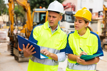 Man and woman builders in uniform and hardhats discussing while standing in construction site. Man using laptop, woman holding folder with documentation.
