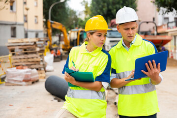 Two civil engineers with laptop and documents checking work process in construction site outdoors