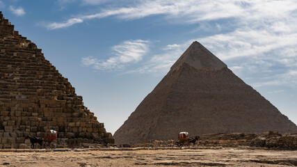 Fototapeta na wymiar Two great pyramids - Cheops and Chephren on a background of blue sky and clouds. Horse-drawn carts are visible at their foot. Egypt. Giza