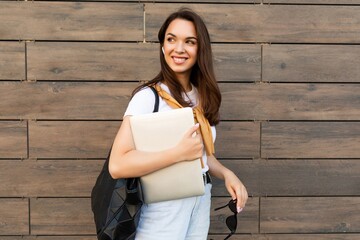 Photo of pretty attractive beautiful smiling happy positive charming young brunette young woman holding gray computer laptop wearing stylish clothes in white t-shirt and light blue jeans in the street