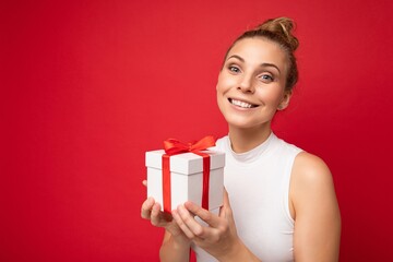 Photo shot of beautiful attractive cute positive happu smiling young blonde woman isolated over red background wall with copy space for text wearing casual white top holding gift box with red ribbon