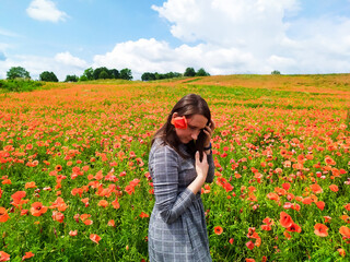 Smiling woman in a long gray dress standing in the middle of the red wildflowers field and cloudy sky background. Happy brunette girl in a red field of poppies stands and enjoys life. Beauty in nature