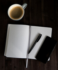 open white notebook , a black mobile phone and a cup of coffee on a dark wooden background