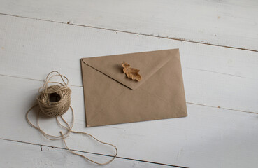 A mock-up of an envelope made of kraft paper, a dried oak leaf and a ball of linen threads on a white wooden surface.