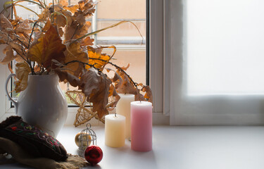 Interior home scene. Ceramic white vase with autumn leaves, candles, a cage, Christmas decorations and a mitten with embroidery on the windowsill against the background of the window.