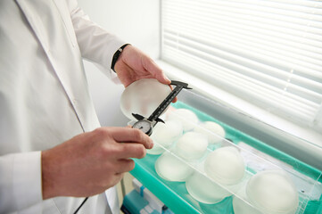 Close-up of hands of plastic surgeon holding a silicone breast implant and measuring its size with...