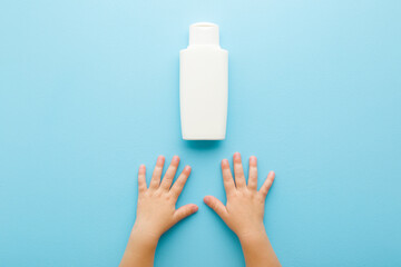Baby hands and white plastic bottle of shampoo or shower gel on light blue table background. Pastel...