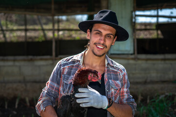 Close-up of Portrait of a cheerful farmer holding and taking care of chicken outdoor. A smart hipster young man holding a hen has been satisfied smiling at an organic sustainable egg farm