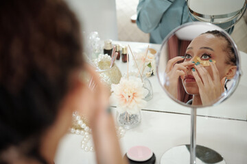 Pretty young woman looking at big mirror and applying anti-wrinkle undereye patches
