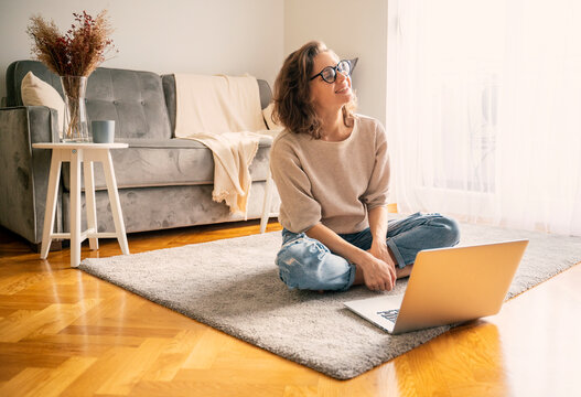 Happy beautiful young woman student sitting at home on carpet in front of laptop