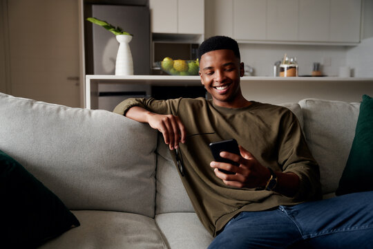 Young Adult Black Male Relaxing On Sofa While Texting In Modern Apartment