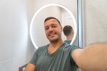 Cheerful handsome unshaven millennial guy taking selfie photo at home with reflection in bahtroom...