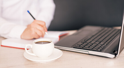 Woman writes with pen in notebook in front of laptop and cup of coffee.