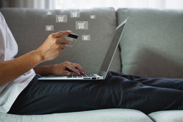 Young man wearing white t-shirt sitting on sofa in living room using laptop and smartphone and hands holding credit card with using shopping cart icon. Show that he is shopping online.