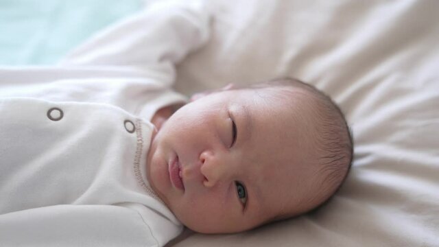 Close up newborn baby in white clothes. Baby in a light blouse wakes up and grimaces.