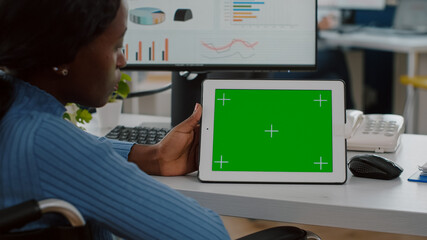 Disabled handicapped african woman employee in wheelchair looking at tablet with green screen chroma key mock up izolated display analysing graphs, financial statistics working in start up office