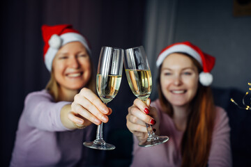 Christmas toast with glasses of champagne in hands. Blurred background: two girls in Santa Claus hats, clink glasses and wish Happy New Year. Love, friendship, family.