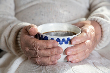 Close-up hands of old woman with mug of hot coffee. Senior lady sitting in chair with blanket. Warm and cozy, time to relax. 