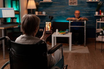 Aged woman holding smartphone for video call conference with relatives while sitting in living room. Elder pensioner talking to daughter and niece with online internet in wheelchair