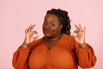 Young black overweight body positive woman shows OK gesture with funny grimace on pink background closeup