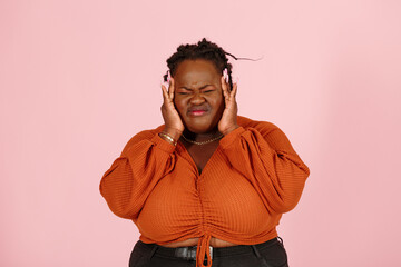 Young black plus size body positive woman patient in orange top suffers from severe headache standing on light pink background in studio closeup