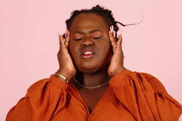 Young black plus size body positive woman patient in orange top suffers from severe headache standing on light pink background in studio closeup