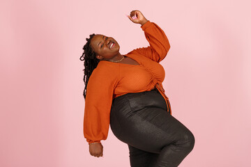 Excited young black plus size body positive woman with dreadlocks in orange top dances standing on light pink background in studio closeup - 468122626