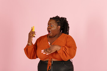 Wondered young black plus size body positive woman with dreadlocks in orange top holds smartphone standing on light pink background in studio closeup