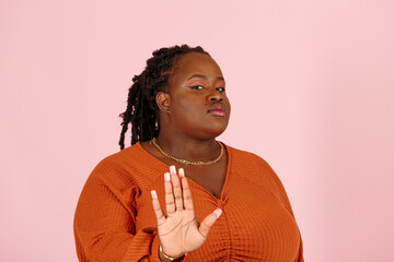Serious young black plus size body positive woman with dreadlocks refuses offer showing Stop gesture on light pink background in studio closeup