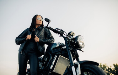 Young woman stands near black motorbike against sky background.