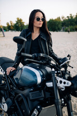 Fototapeta na wymiar Young woman stands near black motorbike among sandy beach.
