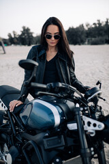 Obraz na płótnie Canvas Young woman stands near black motorbike among sandy beach.