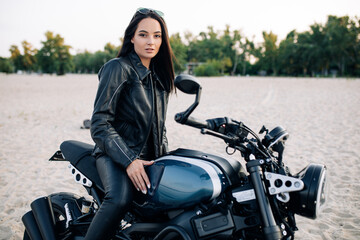 Young woman sits on black motorbike among sandy beach.