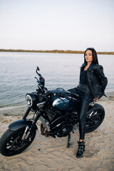 Obraz na płótnie Canvas Young woman sits on black motorbike in beach near river.