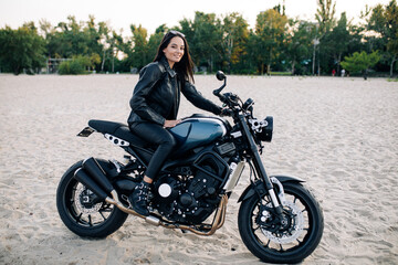 Fototapeta na wymiar Young woman sits on black motorbike among sandy beach.
