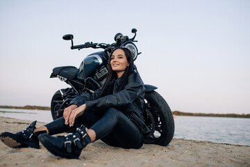 Young woman sits near black motorbike on beach near river.
