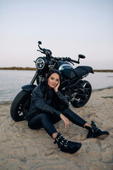 Obraz premium Young woman sits near black motorbike on beach near river.
