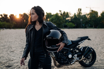 Woman stands with helmet in her hand in beach near motorbike.