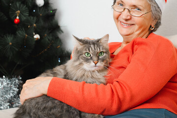 Happy senior woman with cat for Christmas celebrations. Smiling elderly woman in Santa hat sitting with fluffy gray pet alone at home near decorated Christmas tree. Selective focus on cat