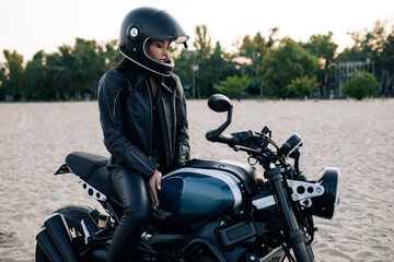 Young woman sits on black motorbike in helmet on beach.