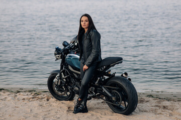 Fototapeta na wymiar Young woman stands next to motorbike on beach near coast of river.