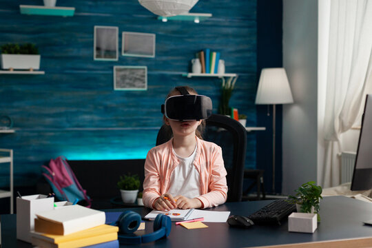 Little schoolkid with virtual reality headset enjoying playing with vr glasses sitting at desk in living room. Child studying literature doing school homework. Home schooling, distance education