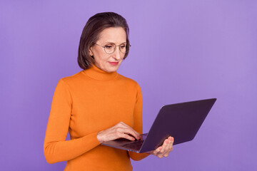 Photo of pretty serious lady pensioner dressed orange turtleneck glasses typing modern device isolated purple color background