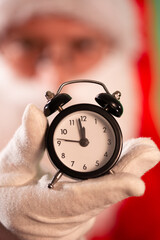 Santa Claus in red coat and hat wearing white gloves showing a small black alarm clock in his hand;...