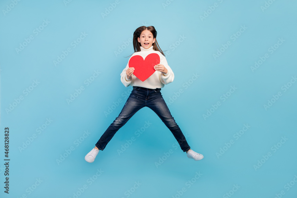 Sticker Photo of adorable impressed student girl wear white sweater smiling jumping high holding large red heart isolated blue color background