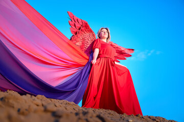 A plump blonde middle-aged woman with red angle wings in a desert with dunes and sand in a nice summer or spring sunny day with blue sky
