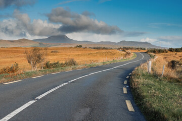 Fototapeta na wymiar Small S shape asphalt road in a country side. Croagh Patrick mountain on the left in clouds. Travel in Ireland. county Mayo. Stunning Irish landscape. Cloud over peak looks like volcano eruption.